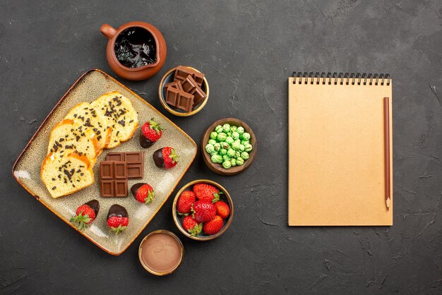 Bovenaanzicht van verre cake en aardbeien kommen van chocolade aardbeien, groene snoepjes en chocoladeroom naast het bord cake en notitieboekje met potlood