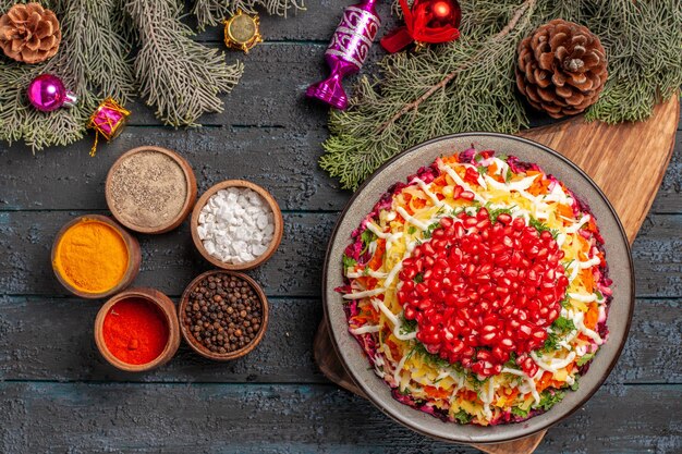 Bovenaanzicht van verre kerstboomspeelgoedschotel met granaatappels op de snijplank kommen met kruiden op het donkere oppervlak naast de boomtakken met kegels en kerstboomspeelgoed