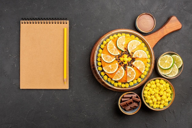 Bovenaanzicht van verre limoenen en snoepjes kommen met verschillende snoepjes schijfjes limoen naast het notitieboekje en potlood en de cake met citrusvruchten op de snijplank op de zwarte tafel