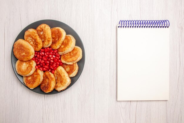 Bovenaanzicht van verre pannenkoeken smakelijke pannenkoeken met granaatappel op de zwarte plaat naast het witte notitieboekje op de witte tafel