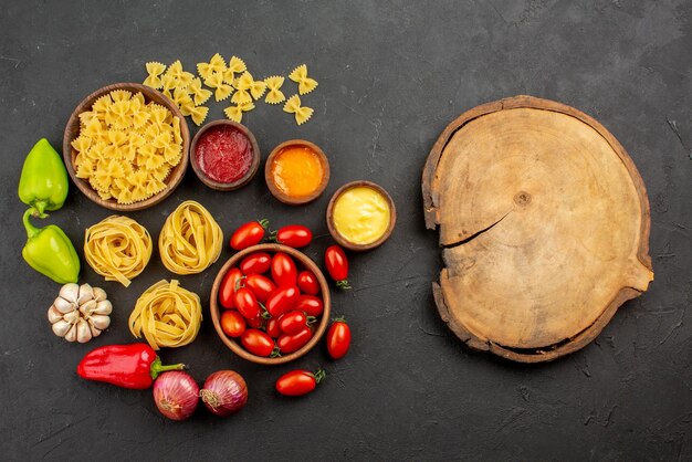 Bovenaanzicht van verre pasta en sauzen tomaten en pasta in kommen paprika knoflook drie soorten sauzen ui naast het houten keukenbord op tafel