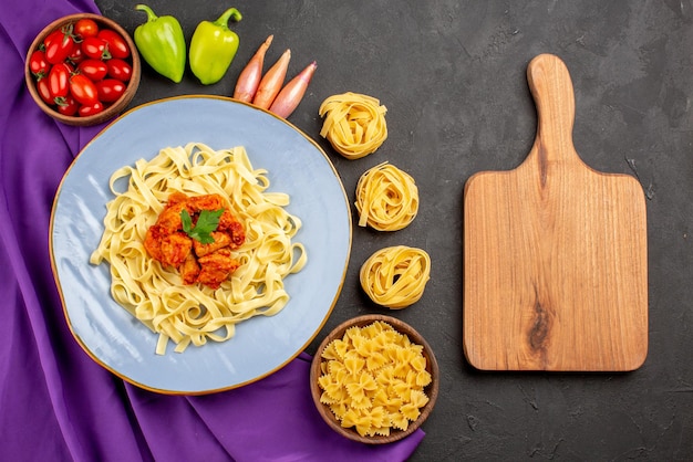 Bovenaanzicht van verre pasta en vleeskom tomaten bal peper ui naast de houten snijplank pasta en bord pasta op het paarse tafelkleed