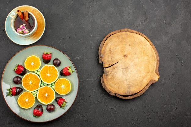 Bovenaanzicht van verre thee met fruit met chocolade bedekte aardbei, smakelijke gehakte sinaasappel en groene snoepjes naast een kopje thee met kaneelstokjes naast snijplank