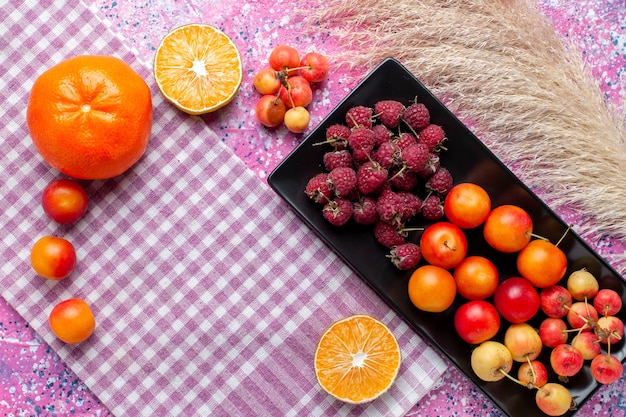 Gratis foto bovenaanzicht van vers fruit, frambozen en pruimen in zwarte vorm met sinaasappels op het roze oppervlak