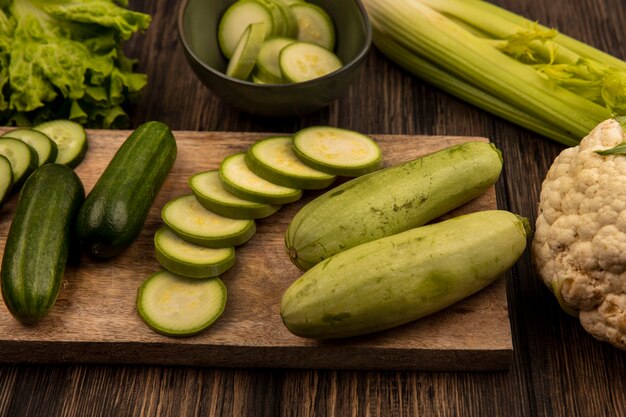 Bovenaanzicht van vers gehakte komkommers en courgettes geïsoleerd op een houten keukenbord met sla selderij en bloemkool geïsoleerd op een houten achtergrond