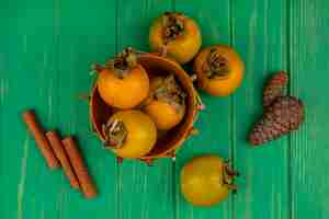 Gratis foto bovenaanzicht van vers kaki fruit op een emmer met kaneelstokjes op een groene houten tafel