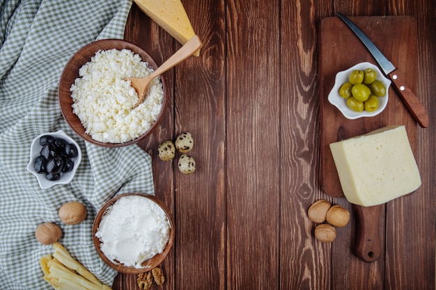 Bovenaanzicht van verschillende kaas en kwark in een kom met walnoten, kwarteleitjes en gepekelde olijven op houten snijplank met een mes op rustieke tafel