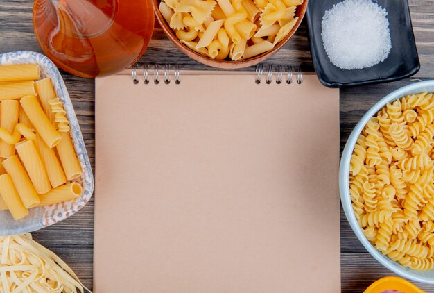 Bovenaanzicht van verschillende soorten pasta als ziti rotini tagliatelle en anderen met gesmolten boterzout rond notitieblok op houten oppervlak met kopie ruimte