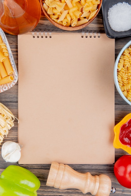 Bovenaanzicht van verschillende soorten pasta als ziti rotini tagliatelle en anderen met knoflook gesmolten boter zout tomatenpeper en ketchup rond notitieblok op houten oppervlak met kopie ruimte