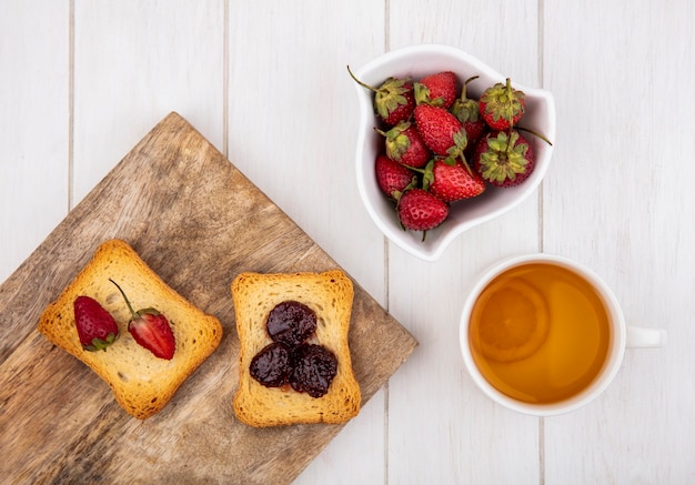 Bovenaanzicht van verse aardbeien op een witte kom met geroosterde sneetjes brood op een houten keukenbord op een witte houten achtergrond