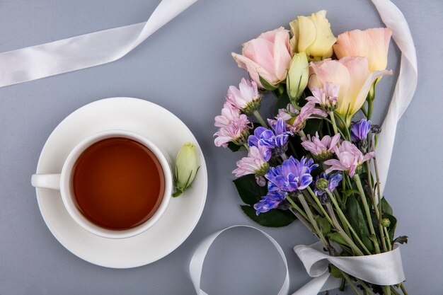 Bovenaanzicht van verse bloemen met wit lint en een kopje thee op een grijze achtergrond