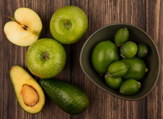 Bovenaanzicht van verse feijoas met limoenen (lemmetjes) op een kom met groene appels en avocado's geïsoleerd op een houten oppervlak