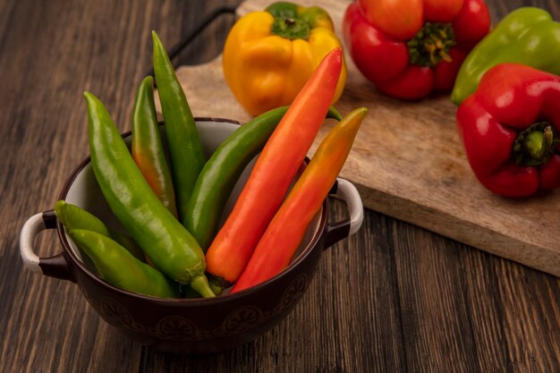 Bovenaanzicht van verse groene en oranje paprika's op een kom met gele rode en groene paprika's op een houten keukenbord op een houten oppervlak