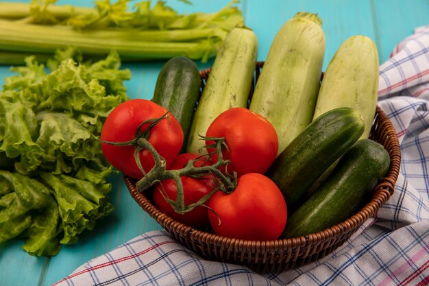Bovenaanzicht van verse groenten zoals tomaten, komkommers en courgettes op een emmer op een geruite doek met selderij en sla geïsoleerd op een blauwe houten ondergrond
