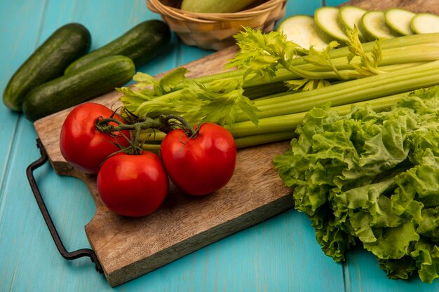 Bovenaanzicht van verse groenten zoals tomaten, selderij en courgettes geïsoleerd op een houten keukenbord met komkommers geïsoleerd op een blauwe houten achtergrond