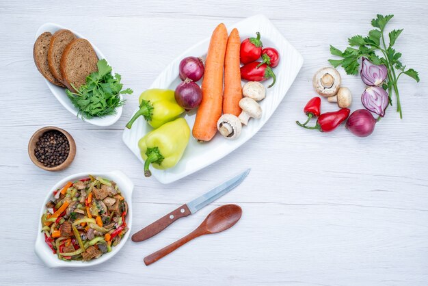 Bovenaanzicht van verse groentesalade gesneden met vlees samen met broodbroodjes en hele groenten en greens op licht bureau, maaltijdsalade vitamine