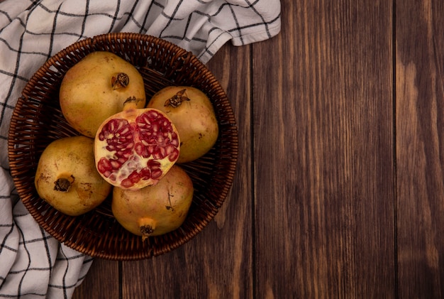 Bovenaanzicht van verse hele en halve granaatappels op een emmer op een gecontroleerde doek op een houten oppervlak met kopie ruimte