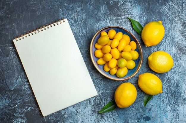 Bovenaanzicht van verse kumquats op een blauw bord en natuurlijke citroenen naast notebook op donkere achtergrond