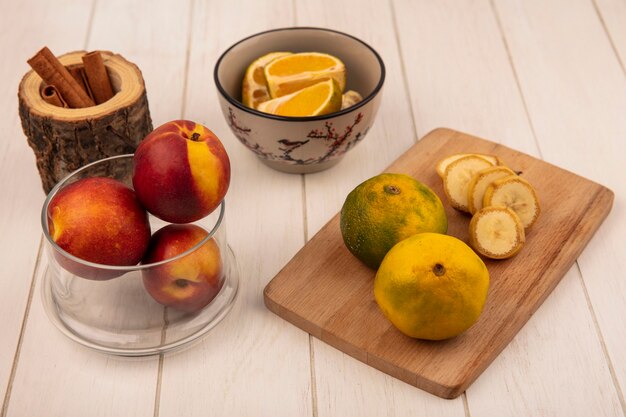 Bovenaanzicht van verse mandarijnen op een houten keukenbord met plakjes banaan met perziken op een glazen kom op een wit houten oppervlak