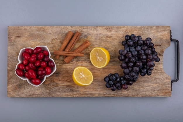 Bovenaanzicht van verse rode cornel bessen op een kom met citroen-kaneelstokjes en druiven op een houten keukenbord op een grijze achtergrond