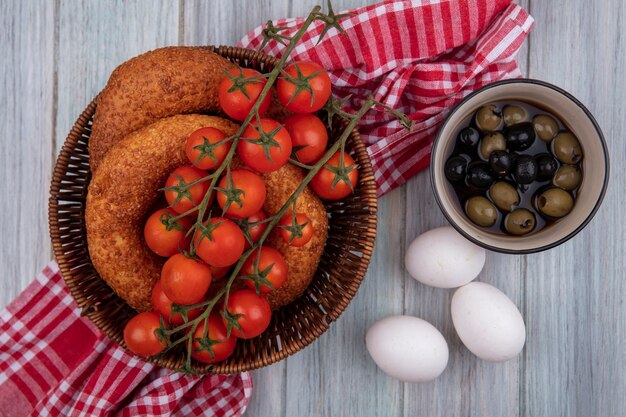 Bovenaanzicht van verse trostomaten op een emmer met bagels op een zakdoek met olijven op een kom en eieren op een grijze houten achtergrond