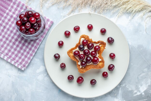 Bovenaanzicht van verse zure kersen in plaat met stervormige romige cake op licht-witte vloer fruit zure bes zomer