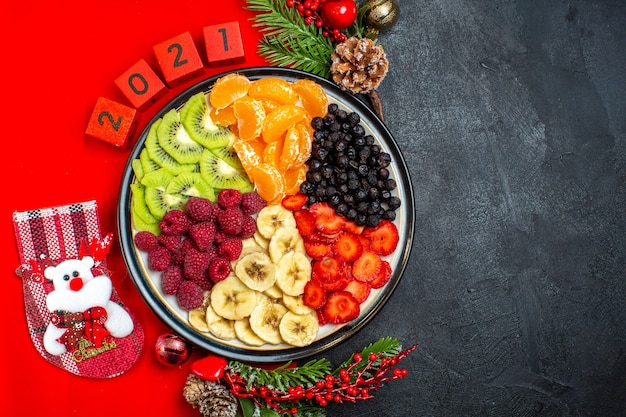 Bovenaanzicht van verzameling van vers fruit op diner plaat decoratie accessoires fir takken en cijfers Kerst Sok op een rode servet op een zwarte achtergrond