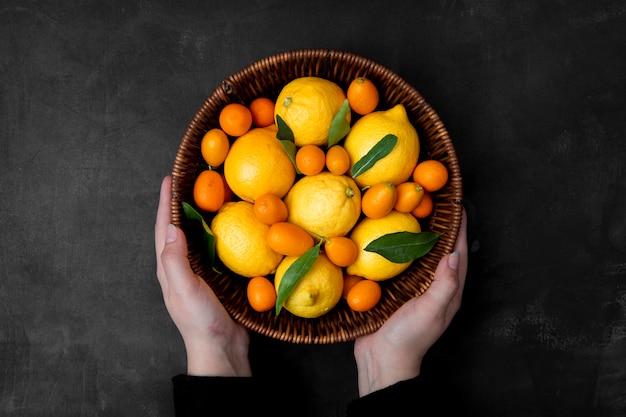 Bovenaanzicht van vrouw handen met mand met citrusvruchten als citroenen en kumquats op zwarte ondergrond