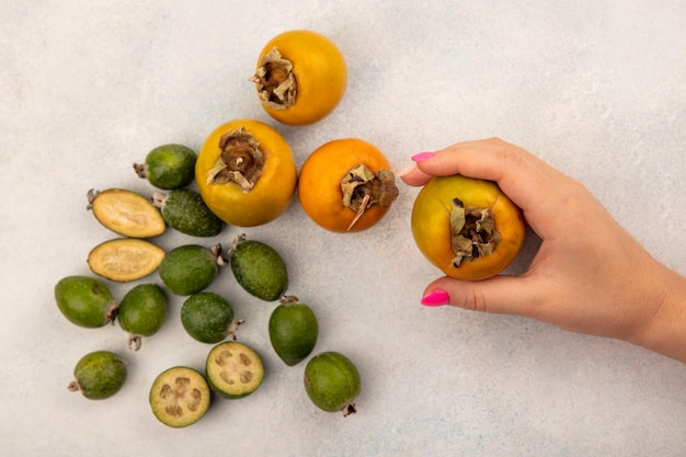 Gratis foto bovenaanzicht van vrouwelijke hand met een biologische kaki fruit met feijoas en kaki geïsoleerd op een grijze ondergrond