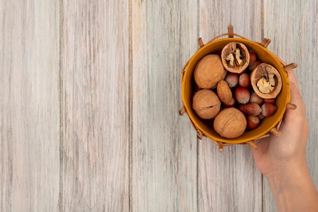 Bovenaanzicht van vrouwelijke hand met een emmer met verschillende noten zoals walnoten en hazelnoten op een grijze houten ondergrond met kopie ruimte