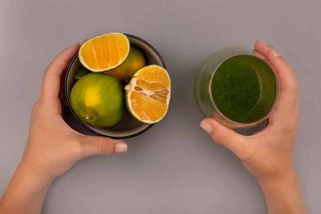 Bovenaanzicht van vrouwelijke hand met een glas vers kiwisap in de ene hand en in de andere hand een kom met mandarijnen