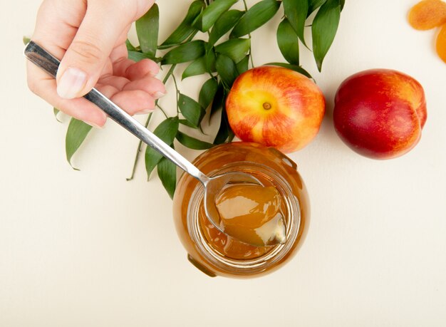 Gratis foto bovenaanzicht van vrouwelijke hand met een lepel met perzik jam over de glazen pot en verse nectarines met groene bladeren op wit