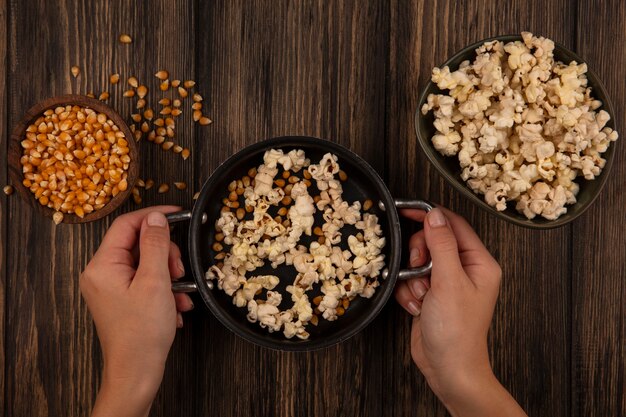 Bovenaanzicht van vrouwelijke hand met een pan met popcorn met maïskorrels op een houten kom op een houten tafel