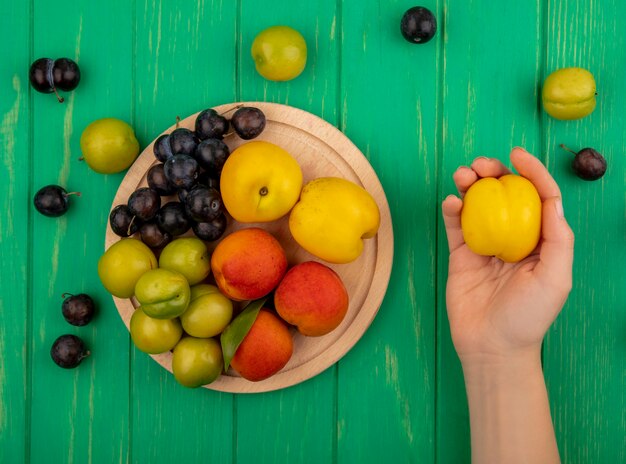 Bovenaanzicht van vrouwelijke hand met gele perzik met vers fruit zoals perzikenloesgroene kersenpruimen op een houten keukenbord op een groene achtergrond