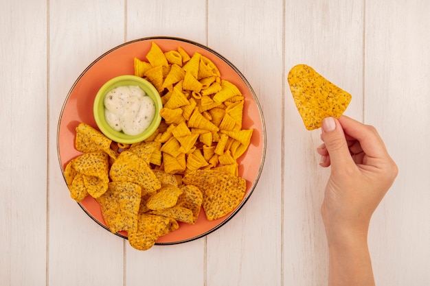 Bovenaanzicht van vrouwelijke hand met pittige knapperige chips met een oranje plaat van pittige knapperige chips met saus op een groene kom op een beige houten tafel