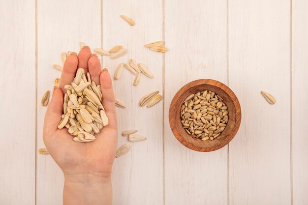 Bovenaanzicht van vrouwelijke hand met smakelijke zoute witte zonnebloempitten met gepelde zonnebloempitten op een houten kom op een beige houten tafel