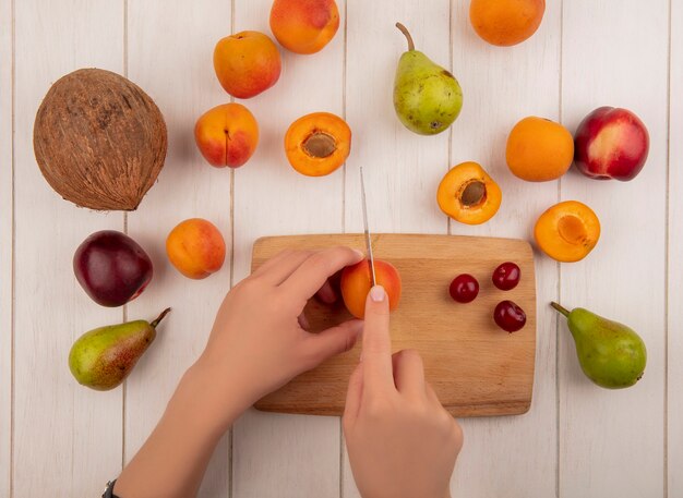 Bovenaanzicht van vrouwelijke handen abrikoos snijden met mes en kersen op snijplank met patroon van peren perzik abrikozen en kokos op houten achtergrond