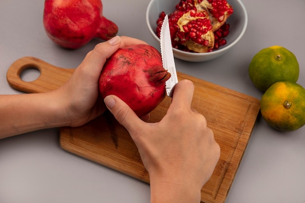 Bovenaanzicht van vrouwelijke handen granaatappels snijden op een houten keuken bord met mes met open granaatappel op een kom met mandarijnen geïsoleerd