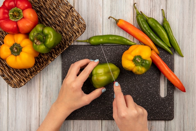 Bovenaanzicht van vrouwelijke handen groene paprika snijden op zwarte keuken bord met mes met paprika geïsoleerd op een grijze houten muur