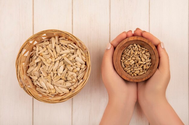 Bovenaanzicht van vrouwelijke handen met een houten kom met smakelijke zoute gepelde zonnebloempitten met zonnebloempitten op een emmer op een beige houten tafel