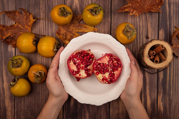 Gratis foto bovenaanzicht van vrouwelijke handen met een kom granaatappels met kaki fruit geïsoleerd op een houten tafel