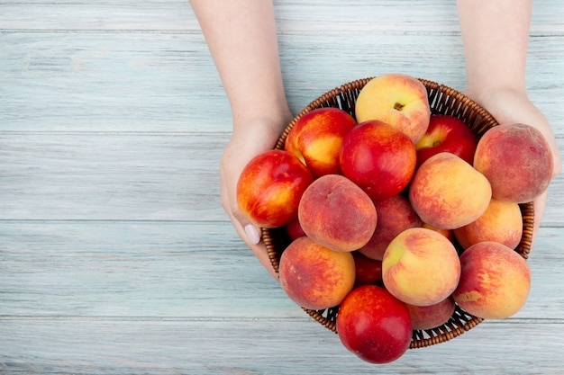 Bovenaanzicht van vrouwelijke handen met een rieten mand met verse rijpe nectarines en perziken op rustiek hout met kopie ruimte