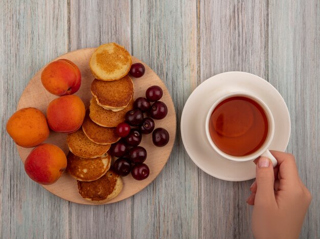 Bovenaanzicht van vrouwelijke handen met kopje thee en pannenkoeken met abrikozen en kersen op snijplank op houten achtergrond