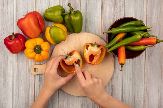 Bovenaanzicht van vrouwelijke handen oranje paprika snijden op een houten keuken bord met mes op een grijze houten oppervlak