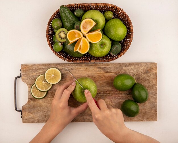 Bovenaanzicht van vrouwelijke handen snijden een appel op een houten keukenbord met mes met een emmer groene appels kiwi feijoas en limoenen (lemmetjes) op een witte muur