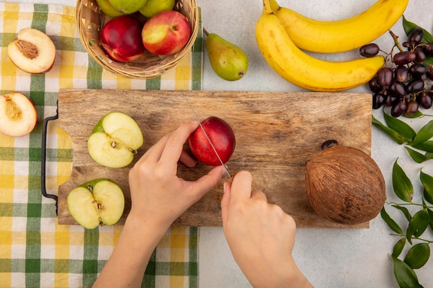 Bovenaanzicht van vrouwelijke handen snijden perzik met mes en half gesneden appel en kokosnoot op snijplank en mandje van perzik appel op geruite doek met bananen druif en bladeren op witte achtergrond