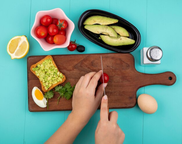 Bovenaanzicht van vrouwelijke handen snijden tomaat met een mes op houten keukenbord met tomaten op kom avocado plakjes en citroen op blauw