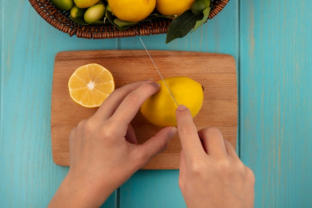 Bovenaanzicht van vrouwelijke handen snijden verse citroen op een houten keukenbord met mes met fruit zoals kinkans en citroenen op een emmer op een blauwe houten ondergrond