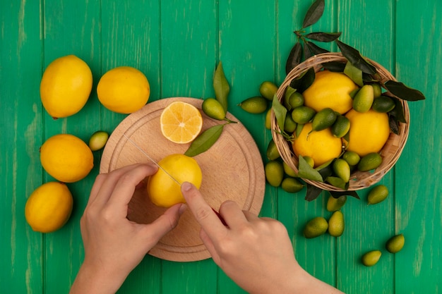 Gratis foto bovenaanzicht van vrouwelijke handen verse citroen snijden op een houten keuken bord met mes met citroenen op een emmer op een groene houten muur