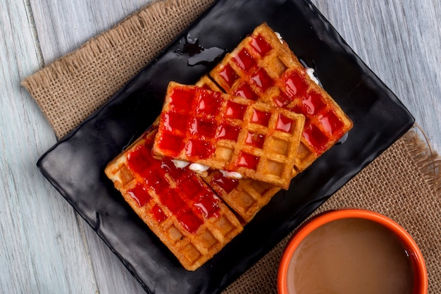 Bovenaanzicht van wafels met aardbeiensiroop en slagroom op een zwarte schotel en een kopje cacao